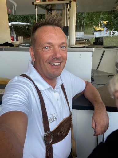 Mann in traditioneller Tracht mit Lächeln, steht vor einem Verkaufsstand.